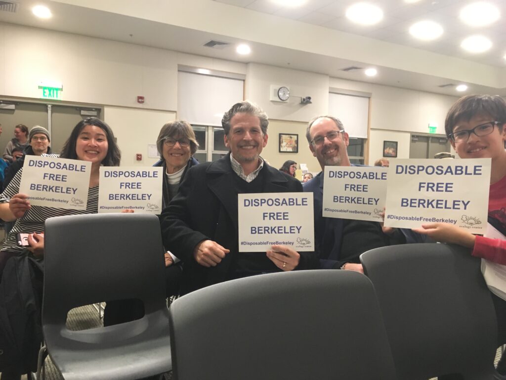 a group of five people sit in Berkeley city hall holding signs that say, "Disposable Free Berkeley"