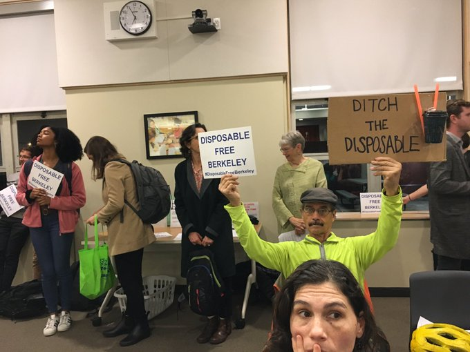 People gathered at a Berkeley city council meeting, a man is holding signs that read, "Disposable Free Berkeley" and "Ditch the Disposables"