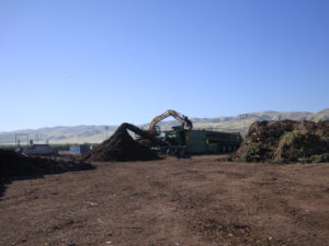Image: A mechanical truck at composting facility working on a pile of compost. There are piles of compost in the foreground and background