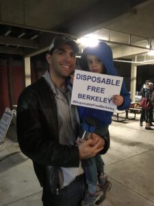 A man holding a child, the child is holding a sign that reads "Disposable Free Berkeley"