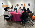 A group of 8 people sit around a table with a magenta table cloth on it. They're having conversations with one another. The table is in a gallery space, with wooden art pieces on the walls.
